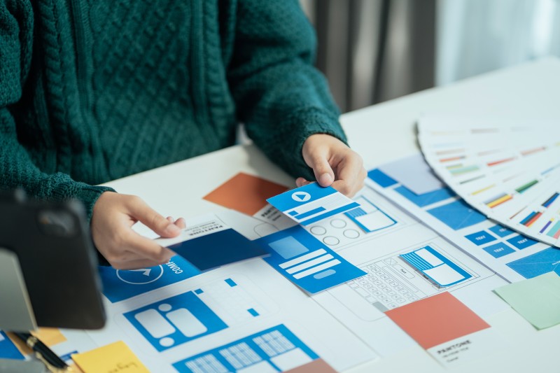 Person's hands in a green sweater arranging blue UI mockup cards and wireframe layouts on a desk beside a colour swatch fan.