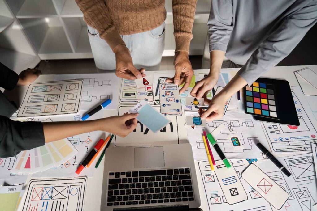 Team collaborating over paper mobile wireframes, placing prototypes, sticky notes, colour swatches, markers and laptop.