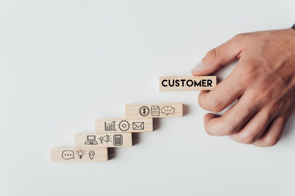 Hand placing a wooden block labeled CUSTOMER atop a staircase of wooden blocks showing business icons.