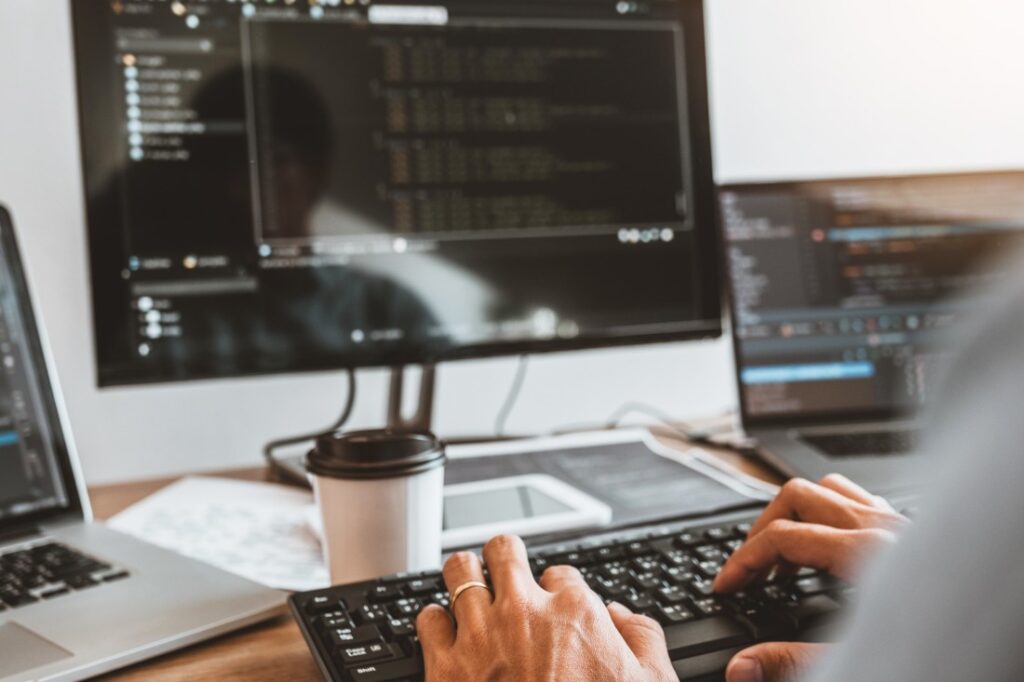 Hands typing on a keyboard at a cluttered desk with code on dual monitors, a laptop and a takeaway coffee cup.