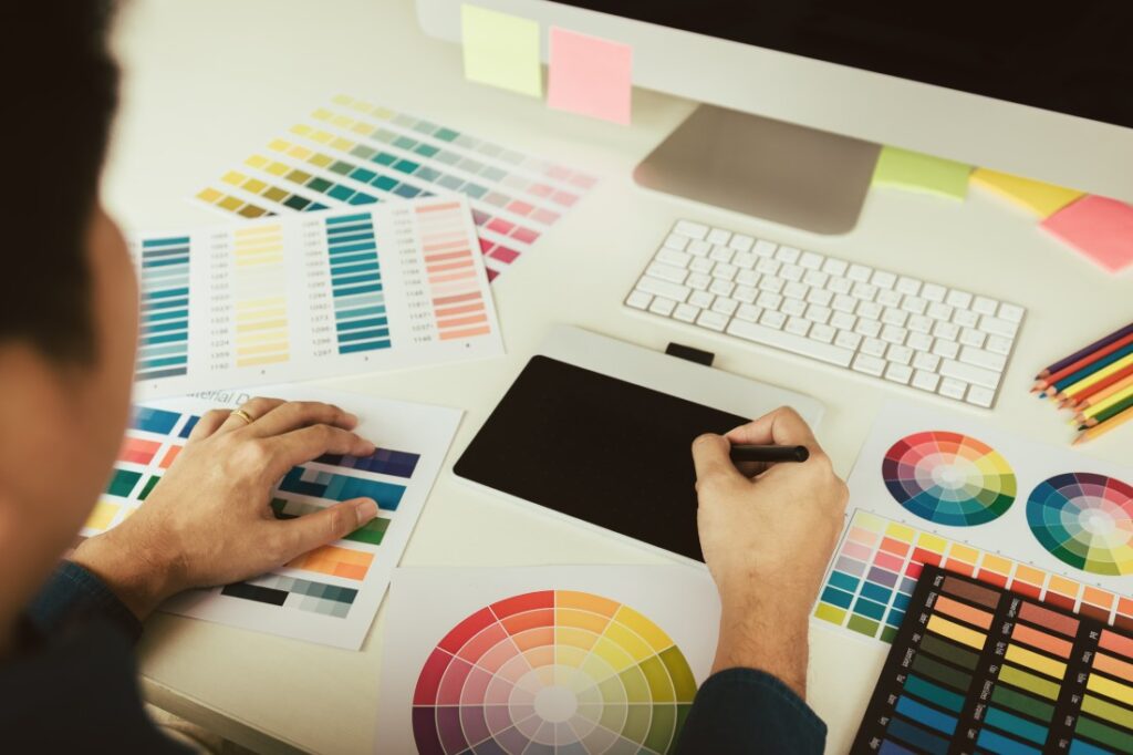Person using a pen tablet while comparing printed colour swatches and colour wheels on a desk
