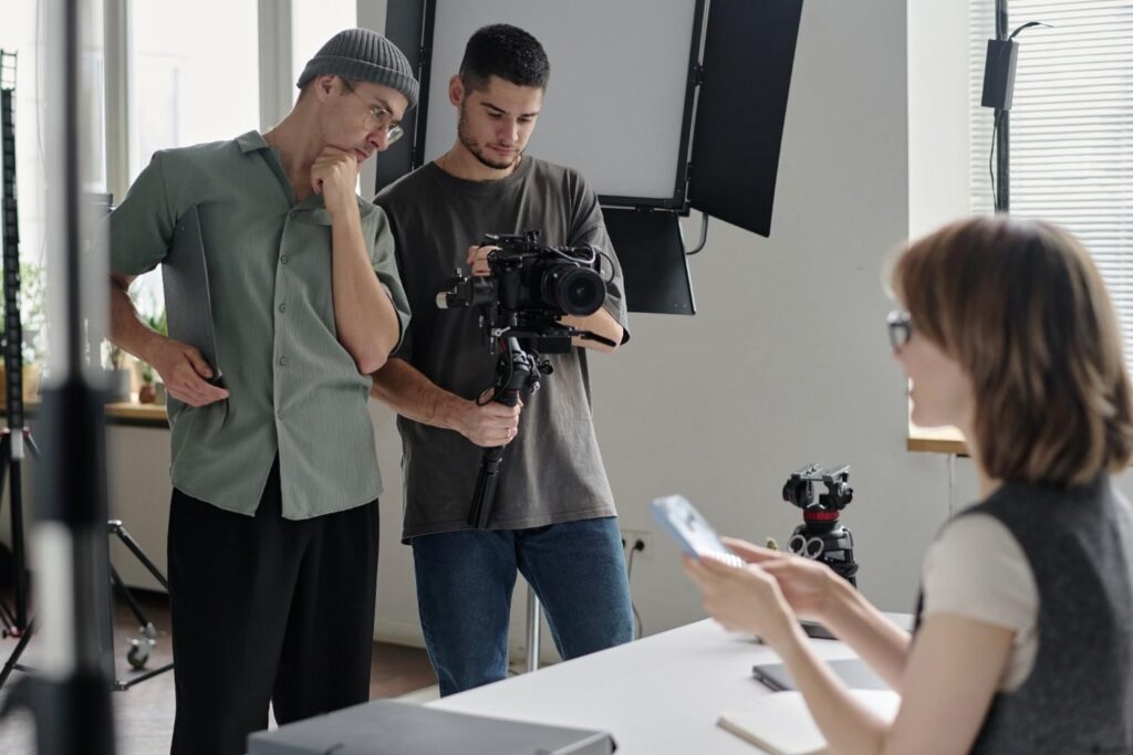 Two men adjust a camera on a stabiliser in a studio while a seated woman in glasses holds her phone in the foreground.