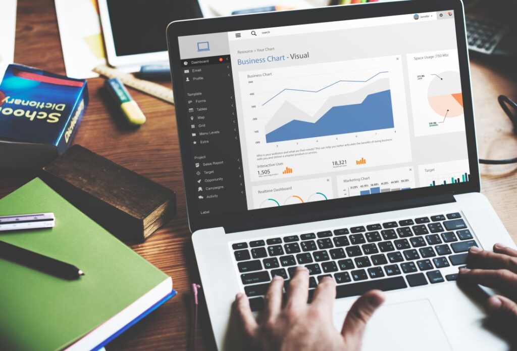 Hands typing on a laptop displaying a business dashboard with line and pie charts on a wooden desk with notebooks