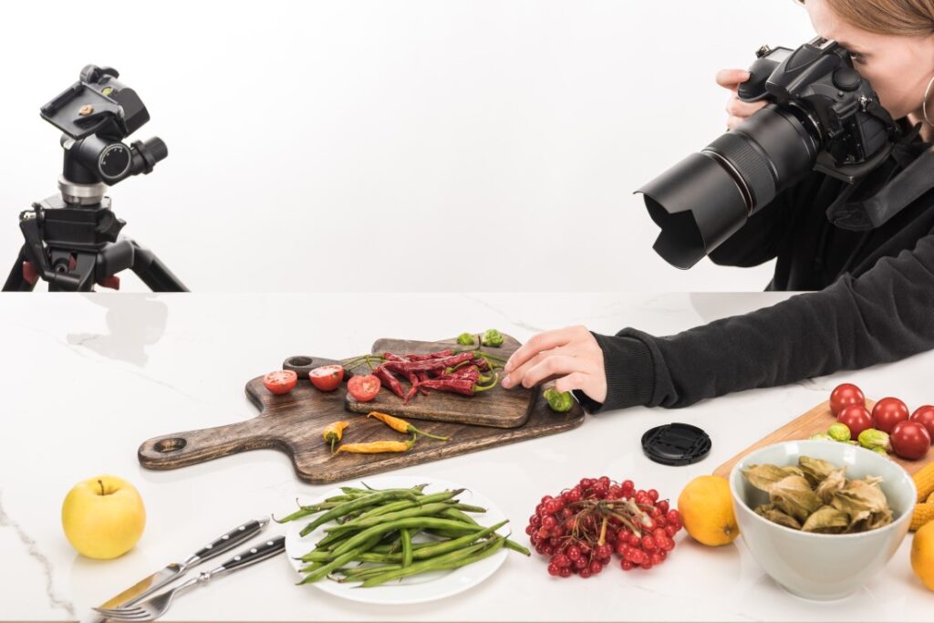 Photographer shooting a food still life: wooden board with red chillies and halved tomatoes, green beans and berries.