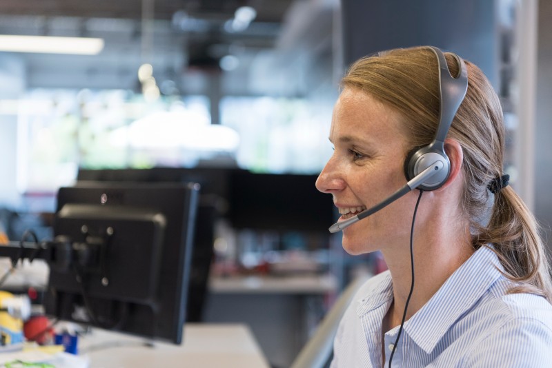 Smiling woman wearing a headset speaking at a computer in a bright open-plan office.