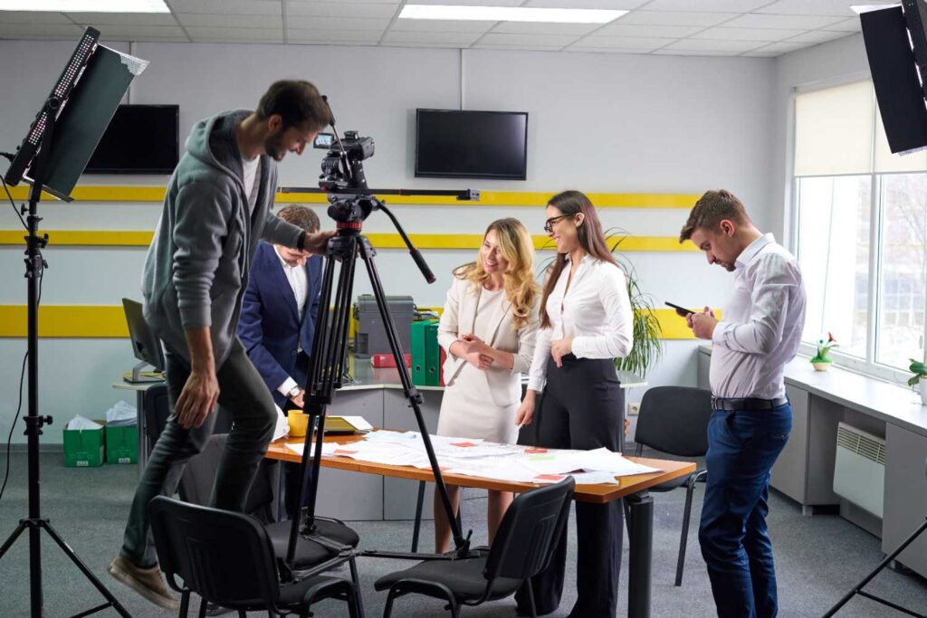 Film crew recording two women discussing documents at an office table, camera on tripod and studio lights set up