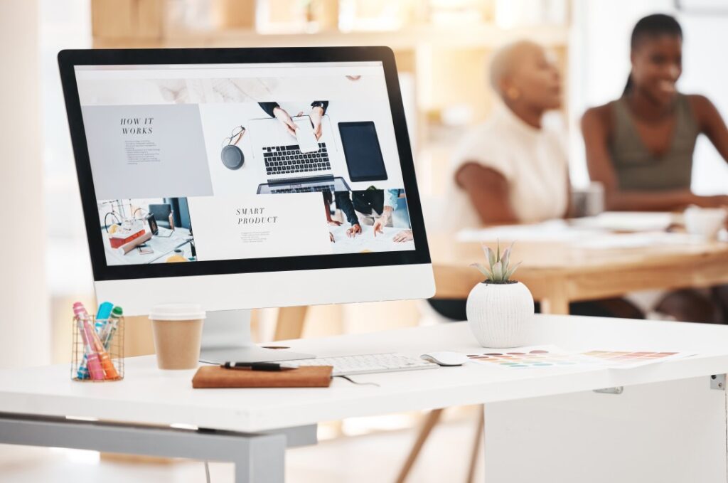 Desktop computer on a white desk displaying a mockup, with coffee, pens, keyboard, succulent and two blurred people.