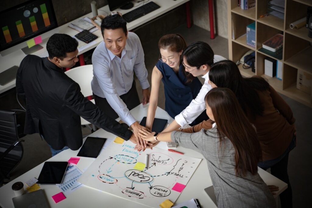 Six colleagues in an office joining hands over a desk with a project diagram, tablets, notes and colourful sticky notes.
