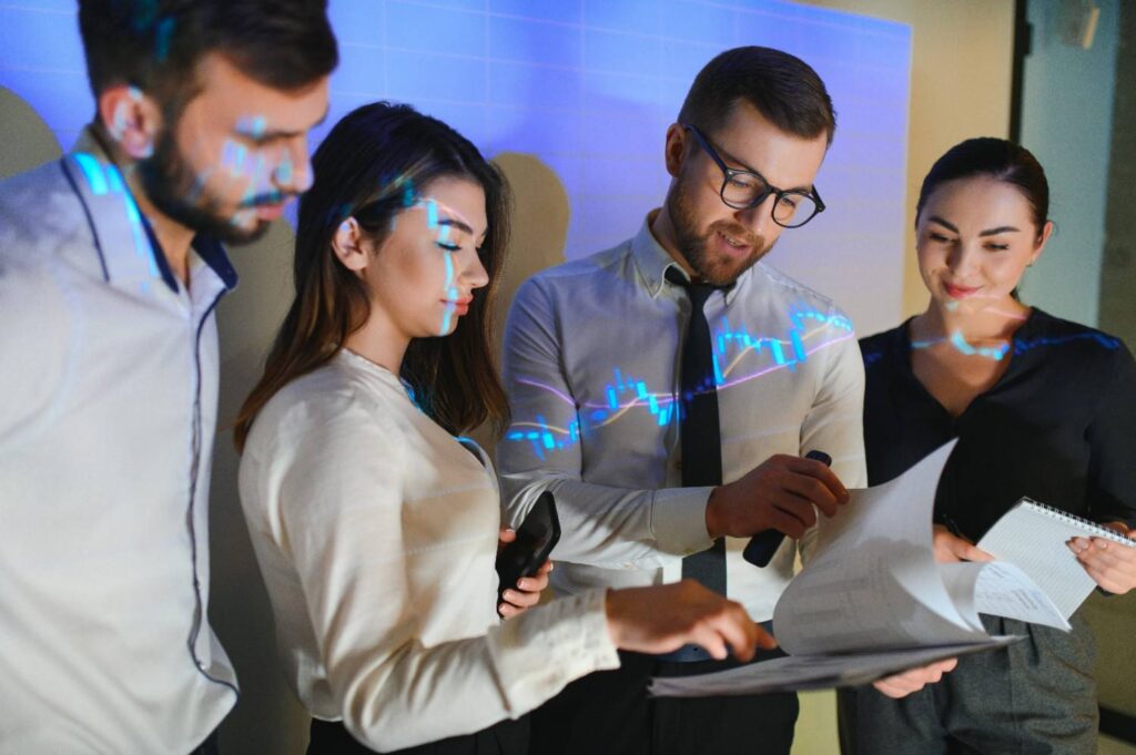 Four colleagues gathered around documents, reviewing papers and notes while a blue stock-chart projection overlays them.