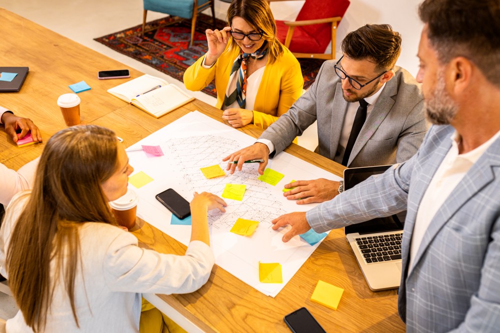 Five colleagues gathered around a table examining a large blueprint covered with colourful sticky notes, laptops and coffee.