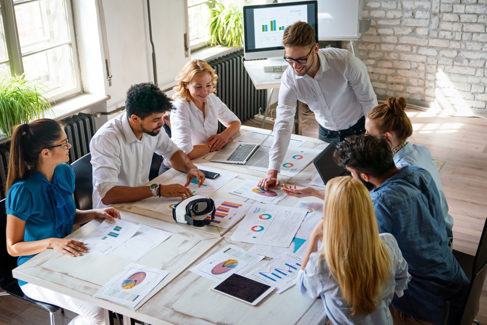 Team of seven colleagues around a table reviewing printed charts, laptops and a tablet during a meeting.