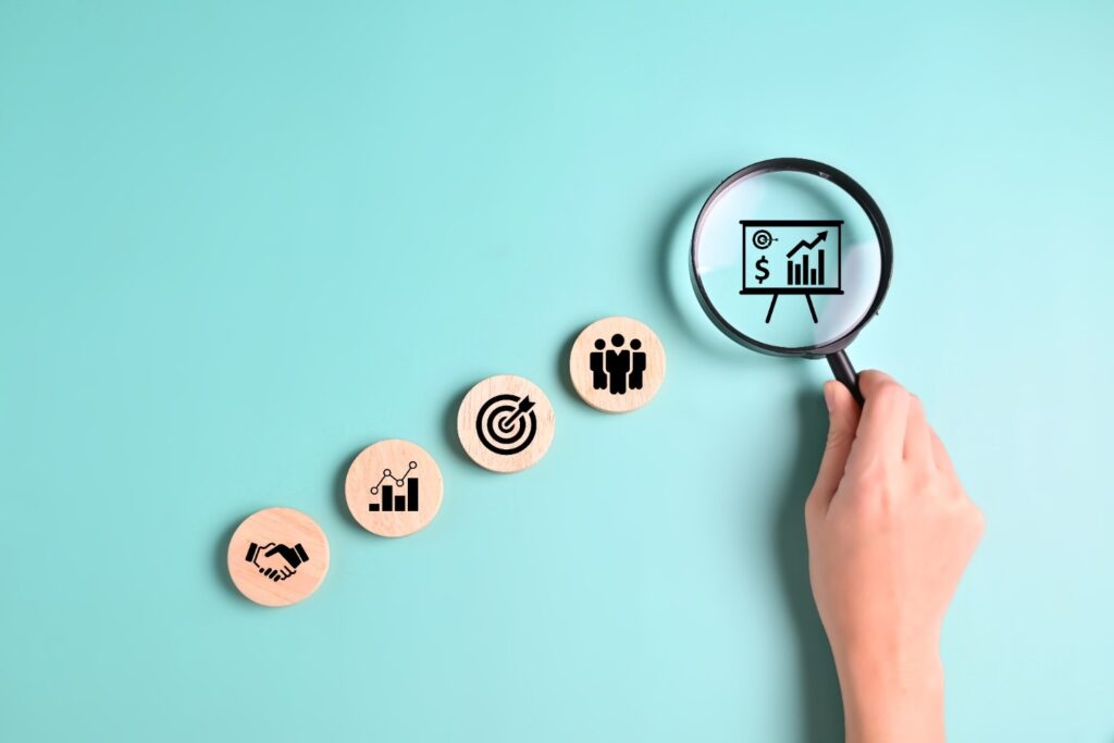 Magnifying glass over a wooden token with rising-chart icon, lined with other wooden business icons on a teal background.