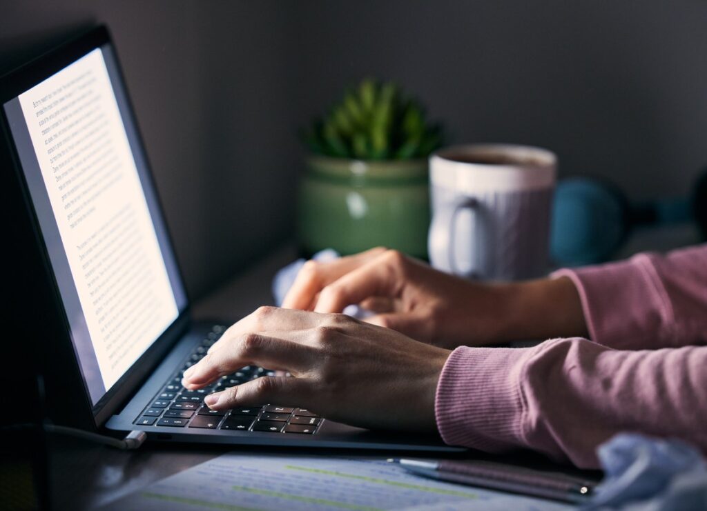 Hands typing on a laptop keyboard beside a mug, potted plant and highlighted notes.