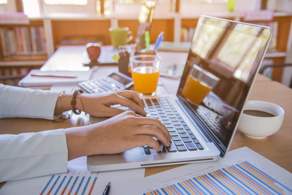 Person's hands typing on a laptop at a cluttered desk with charts, a coffee cup and a glass of orange juice.
