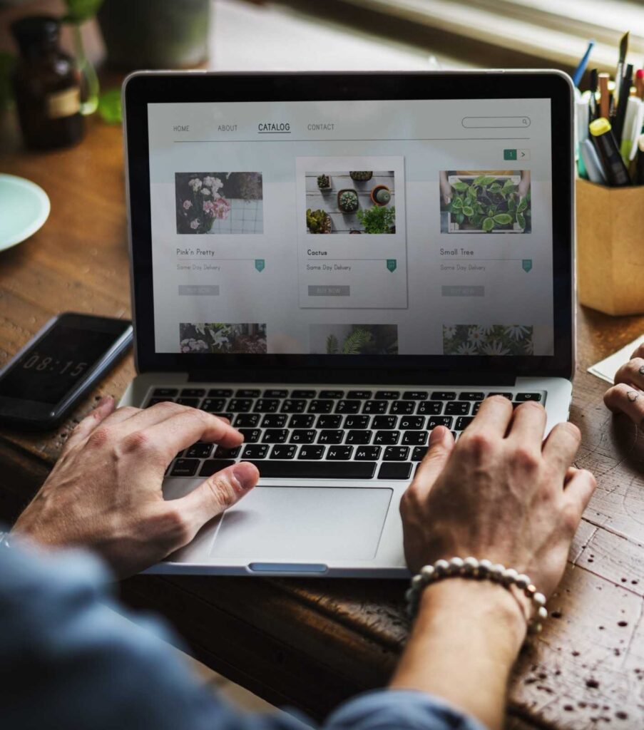 Hands typing on a laptop displaying an online plant catalog with thumbnail photos of potted plants on a wooden desk.