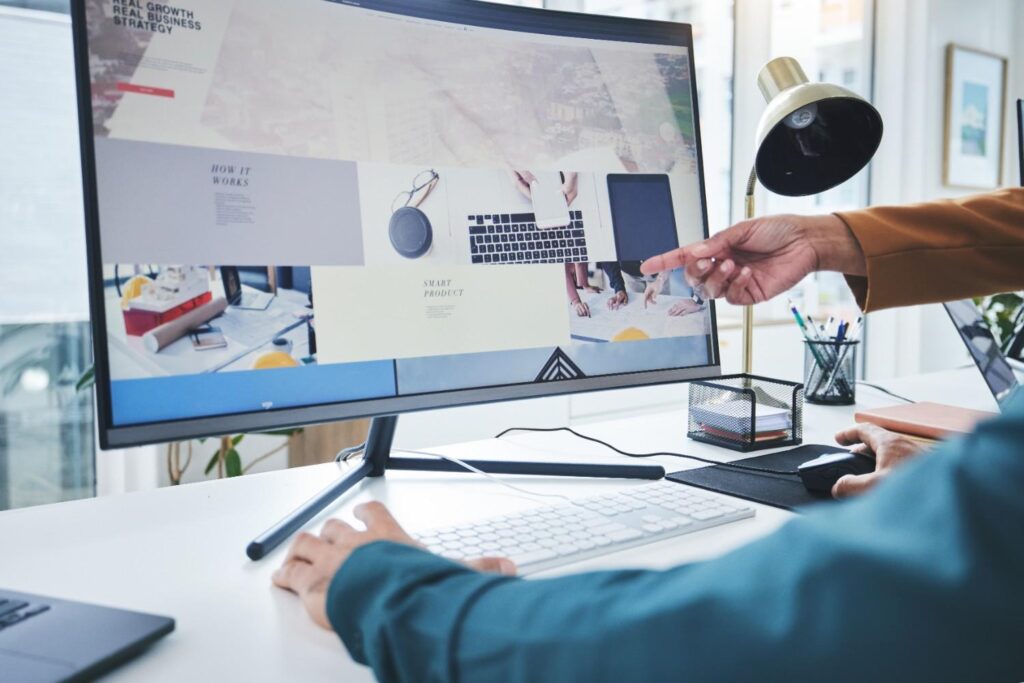Two colleagues reviewing a website mockup on a large desktop monitor, one pointing at the screen while another uses the mouse and keyboard.