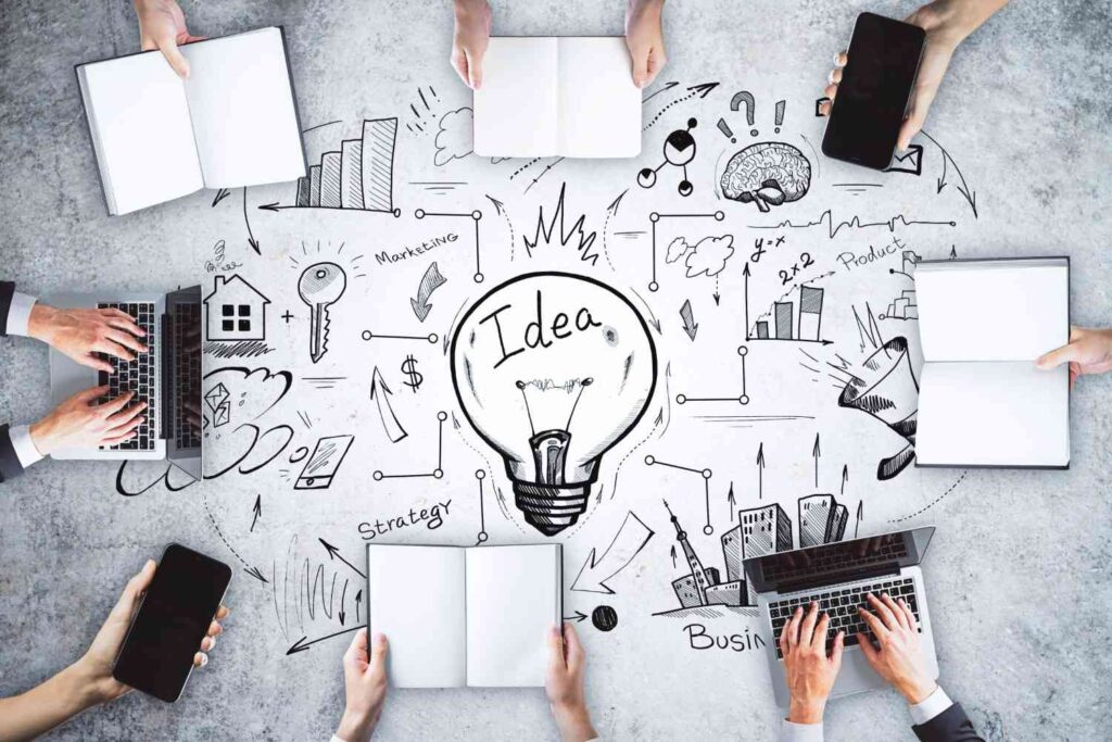Overhead view of people with laptops, phones and notebooks brainstorming around a sketched light bulb labelled Idea.