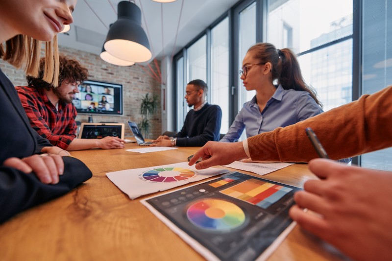 Colleagues around a meeting table examining printed and tablet colour wheels and charts as a laptop displays a video call.
