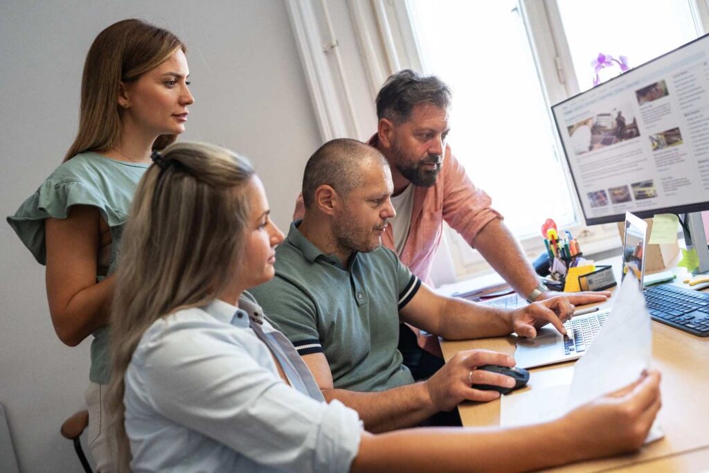 Four coworkers gathered around a desk reviewing documents and a laptop, one man pointing at the screen while others watch.