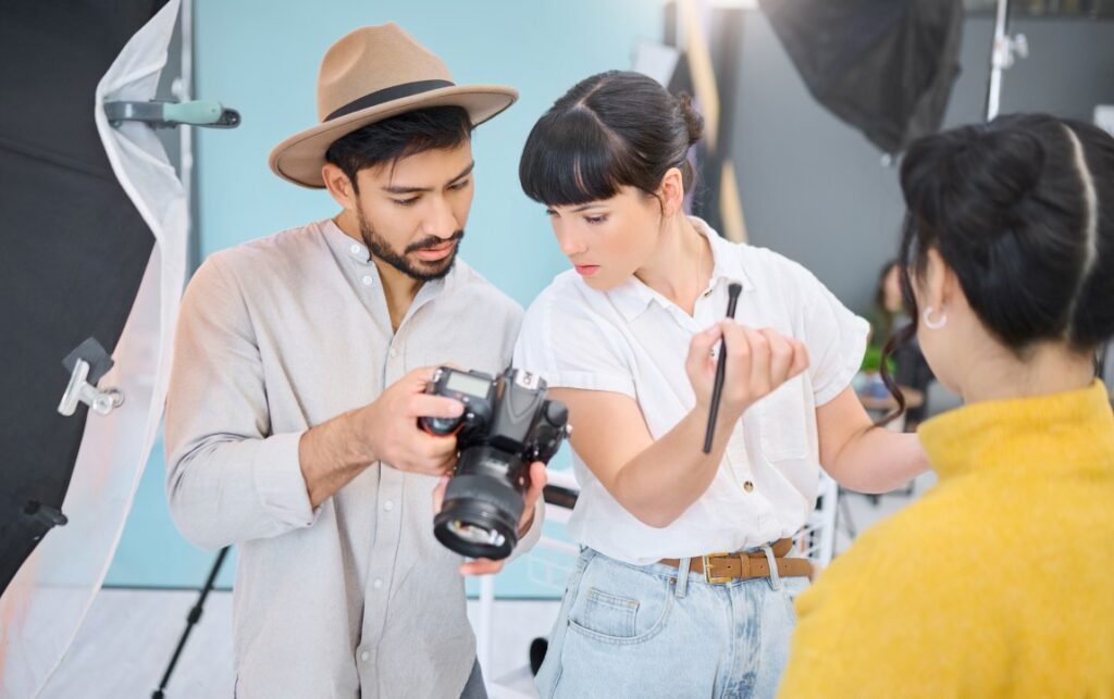 Photographer and stylist check camera images in a studio while a model in a yellow sweater watches.