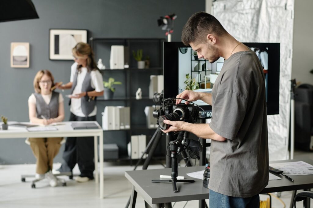 Man adjusting a professional camera on a tripod in a studio, with two blurred women conversing at a desk in the background.
