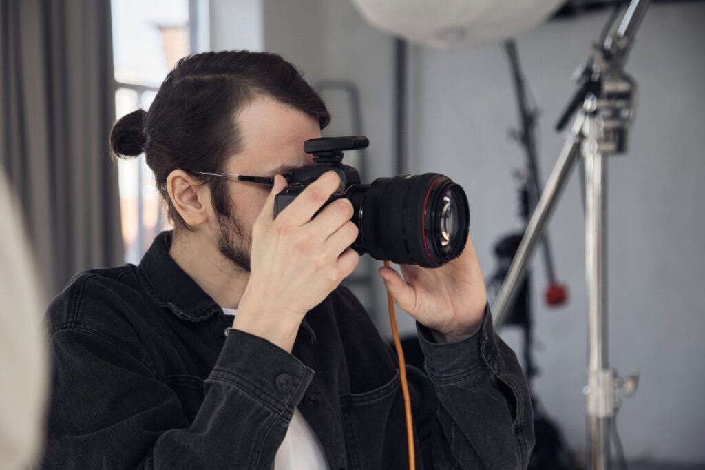 Photographer with a bun and beard wearing glasses holds a DSLR to his eye in a studio with lighting gear.
