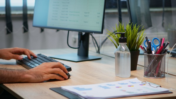 Hands typing on keyboard at office desk with monitor, hand sanitiser bottle, potted plant and paperwork with charts.