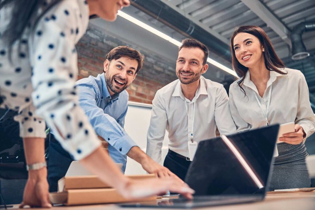 Four colleagues leaning over a laptop, smiling and collaborating around a table in a modern office.