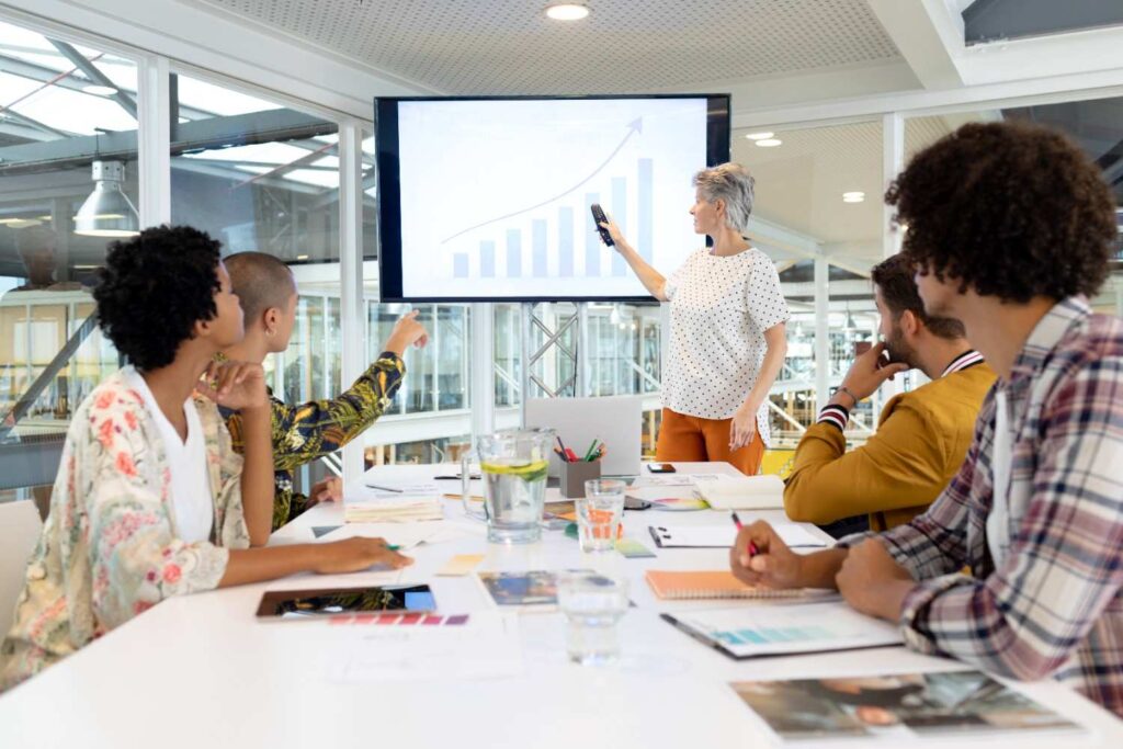 Senior woman pointing at rising bar chart on screen while diverse team around conference table discusses notes and charts