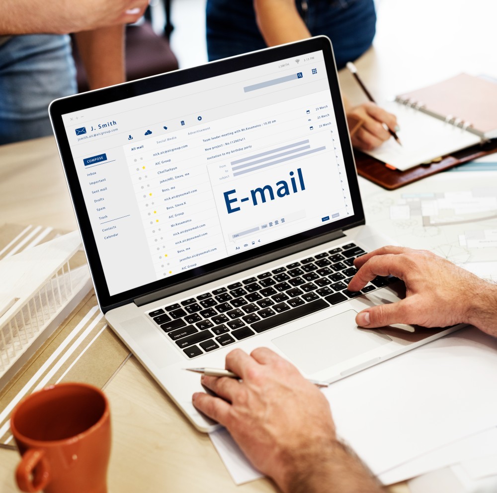 Hands typing on a laptop displaying an open email inbox with the word E-mail on screen, a pen, notebook and coffee cup on the desk.