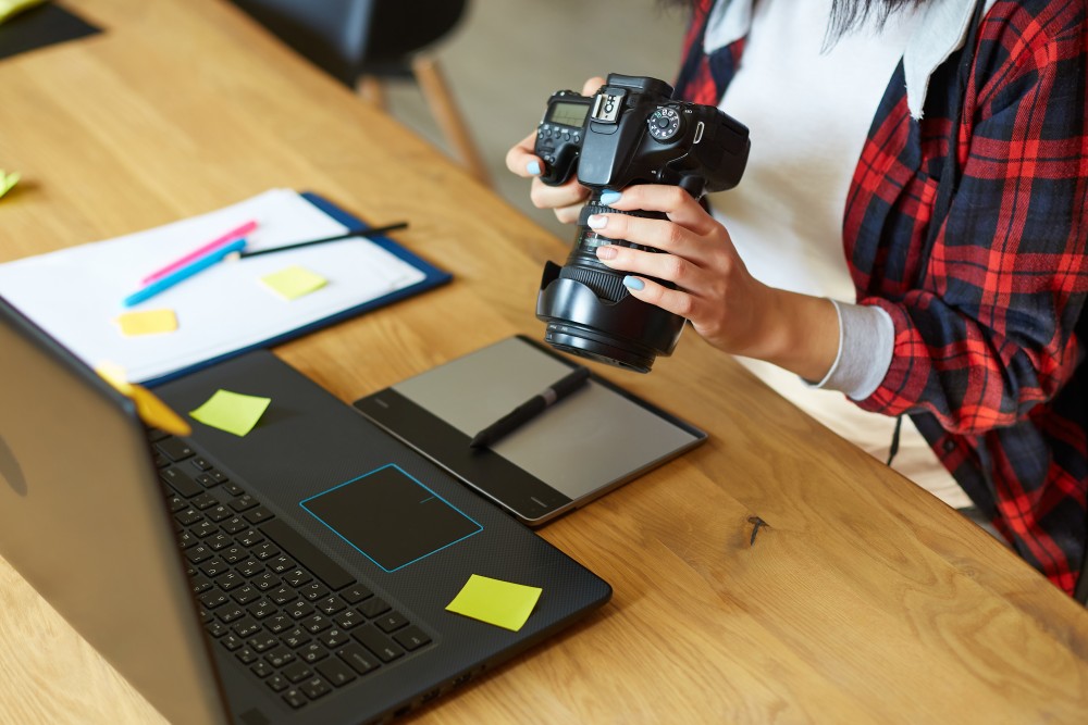 Person in red checked shirt holding a DSLR camera over a laptop and graphics tablet on a wooden desk with sticky notes.