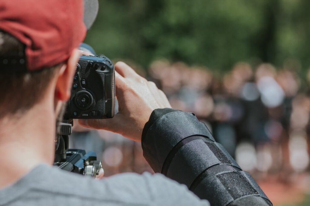 Person in red cap holding a DSLR on a rig, hand on controls and wearing a black forearm brace, photographing a blurred crowd.