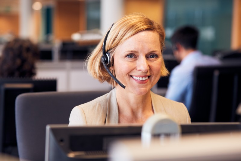 Smiling woman wearing a headset at a busy office customer service desk, computer screens and colleagues blurred behind her.
