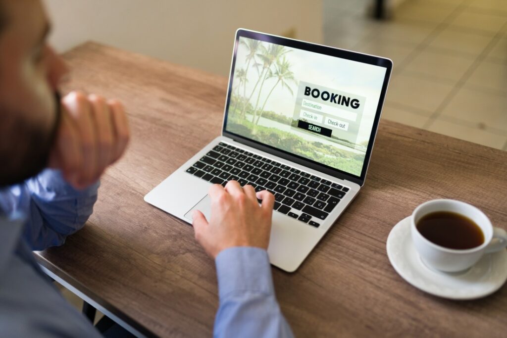 Person using a laptop at a wooden table viewing an online booking page with palm trees on screen and a cup of coffee beside.