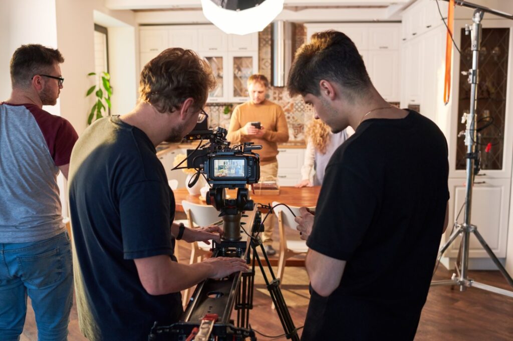Camera crew adjusting a professional camera on a slider in a kitchen set while actors wait under studio lights.