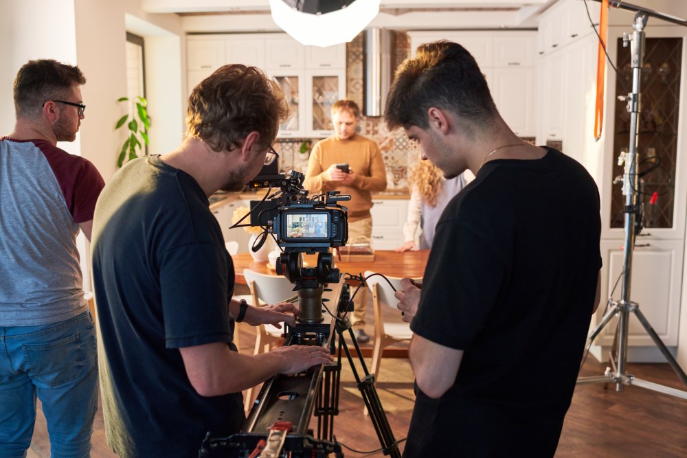 Crew operating a camera on a tripod and slider in a kitchen set, with lighting gear and actors in the background.