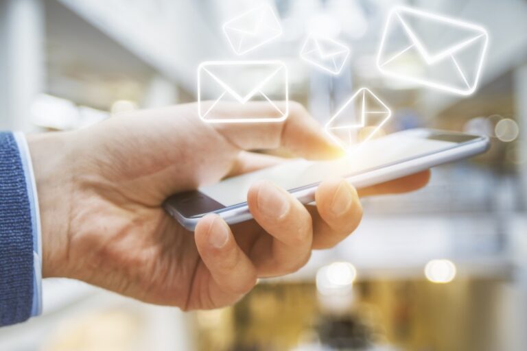 Hand holding a smartphone with glowing envelope icons floating above the screen against a blurred indoor background.