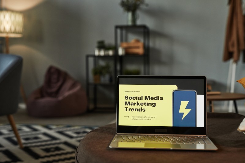 Laptop on an ottoman in a living room showing a yellow slide titled Social Media Marketing Trends with a blue phone icon.