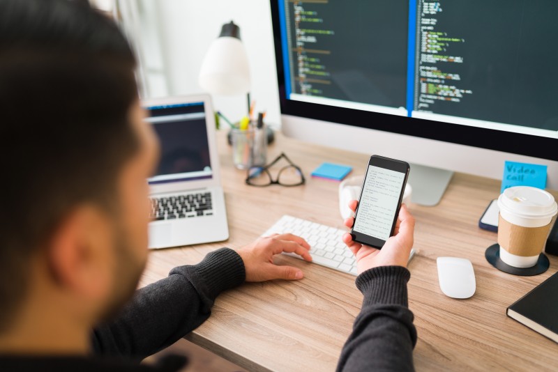 Person at a desk holding a smartphone while looking at code on a desktop monitor, with laptop, keyboard and coffee nearby.
