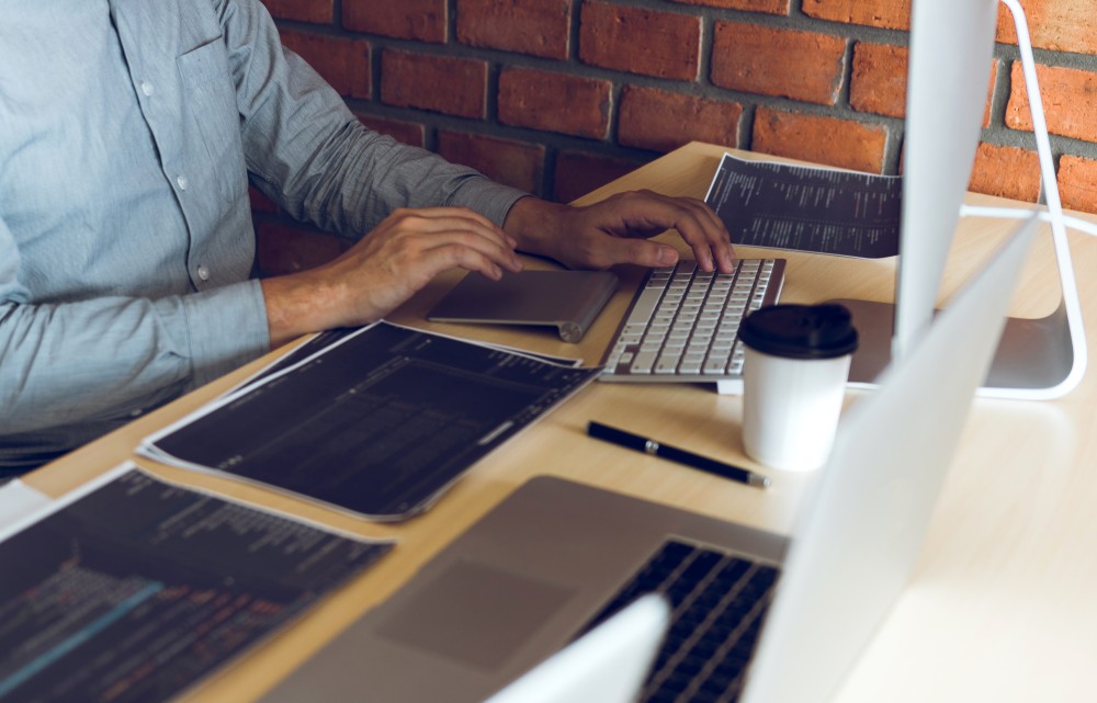 Hands typing on a desktop keyboard surrounded by printed code sheets, a laptop and takeaway coffee against a brick wall.