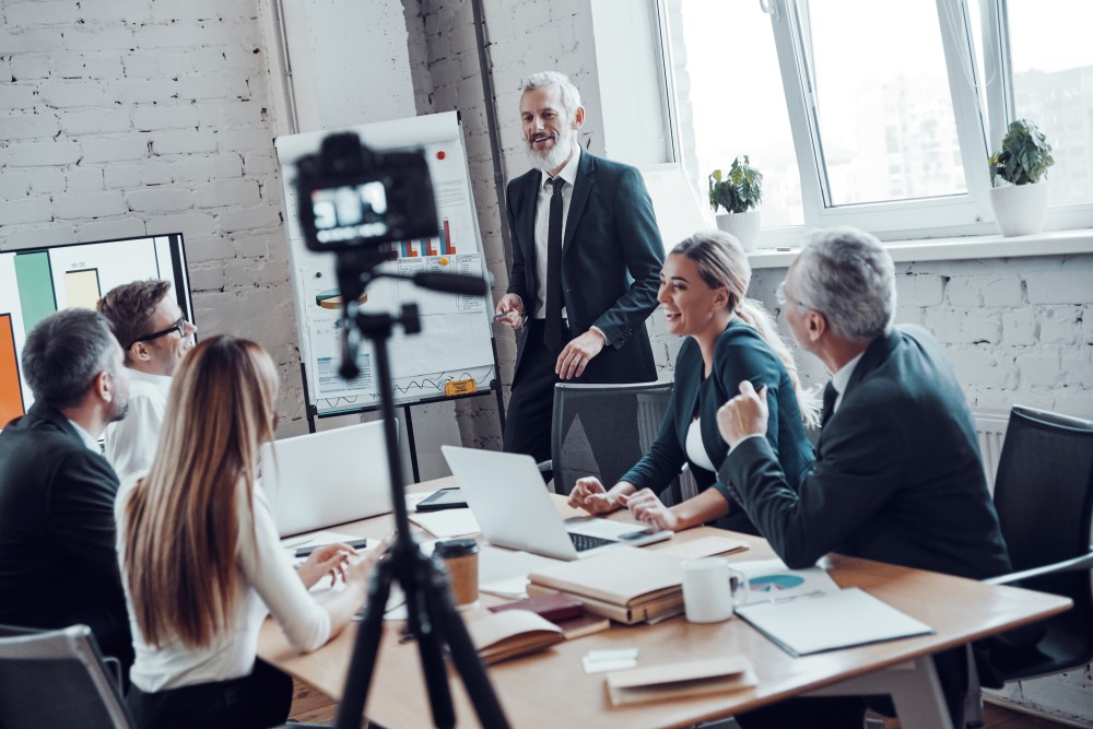 Senior man in suit presenting at a flipchart to smiling colleagues around a conference table, camera tripod in foreground.