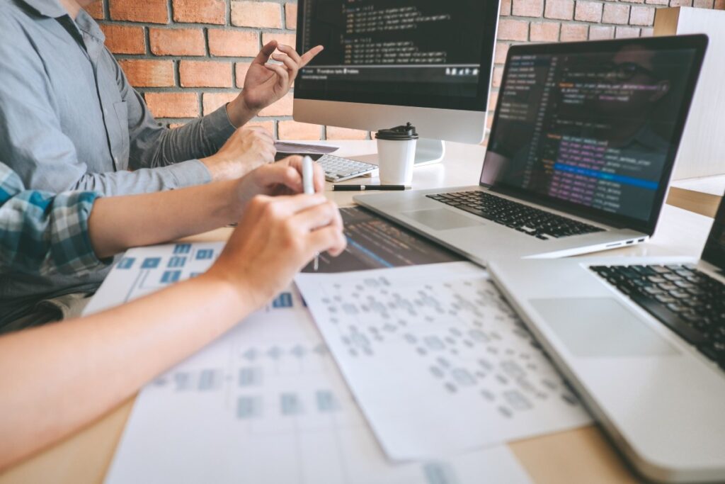 Two developers reviewing code on laptops and monitor, with printed flowcharts and a takeaway coffee cup on the desk.
