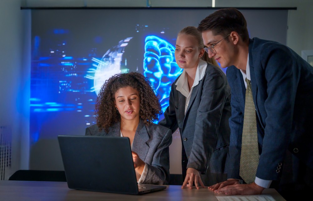 Three colleagues around a laptop, reviewing data while a glowing blue digital brain is projected behind them.