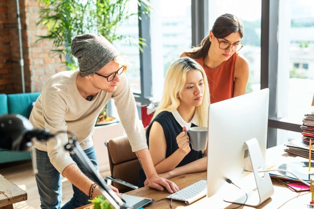 Three coworkers gathered around a desktop: a blonde woman with a mug, a man in a beanie, and a woman with glasses.