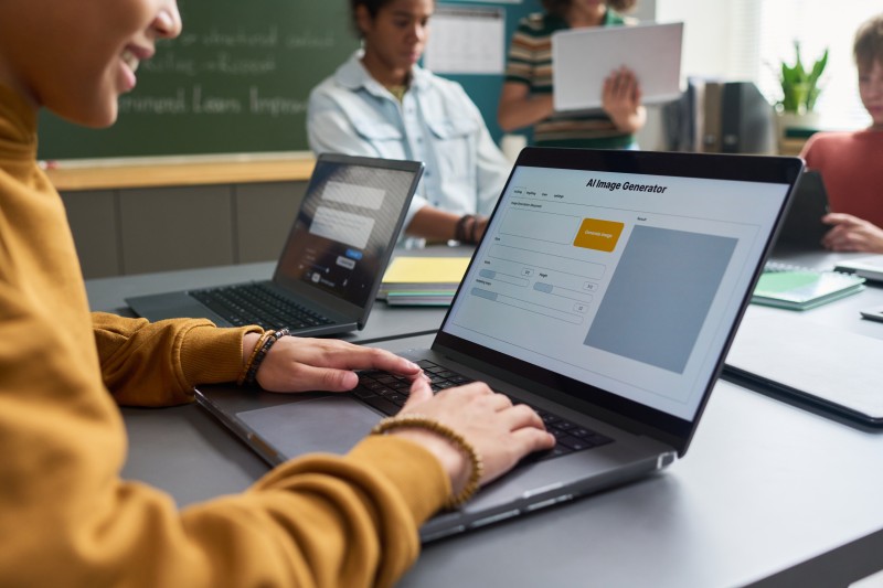 Student typing on a laptop showing an AI Image Generator interface, classmates with laptops in a classroom.