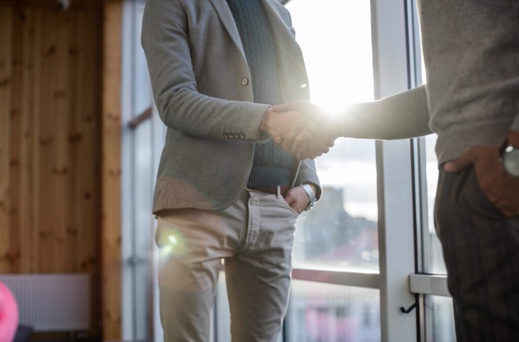 Two people in smart-casual clothing shaking hands by a sunlit window, one with a hand in a pocket.