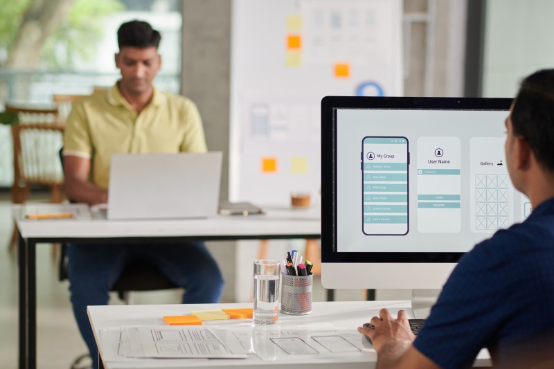 Person at desk working on mobile app wireframes displayed on a monitor, pens and glass on desk, colleague on laptop behind.