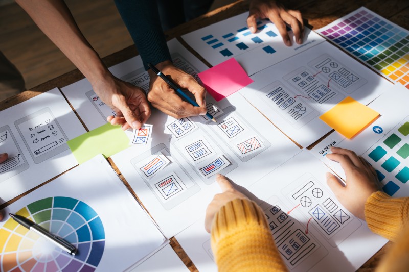Several hands arranging website wireframe sketches, sticky notes and colour swatches spread across a table.