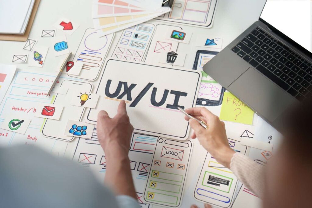 Two people arranging paper UX/UI wireframes and icon sticky notes around a sign reading UX/UI beside a laptop.