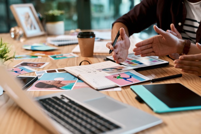 Two people gesturing over a desk strewn with a laptop, notebook, colour swatches, printed photos, tablet and takeaway coffee.